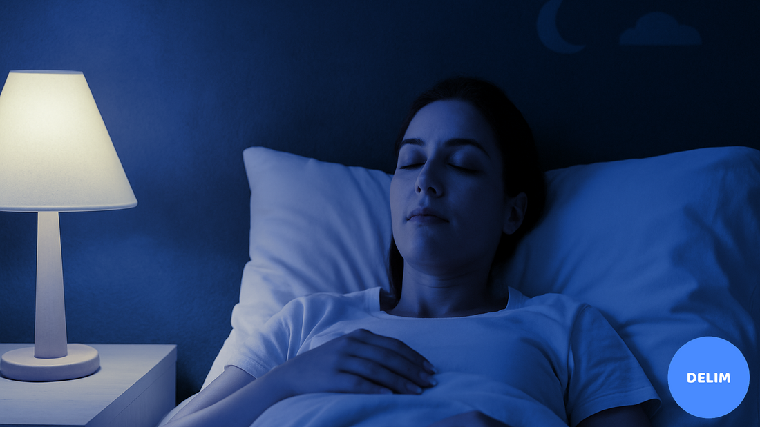 A woman lying peacefully in bed with her eyes closed, dressed in a white T-shirt, illuminated by a warm bedside lamp in a dimly lit bedroom with a dark blue wall.